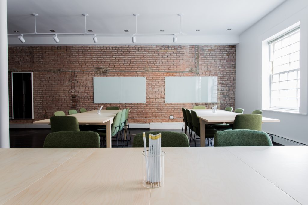Looking over a table in a room with long tables and chairs towards a brick wall with two whiteboards for training.