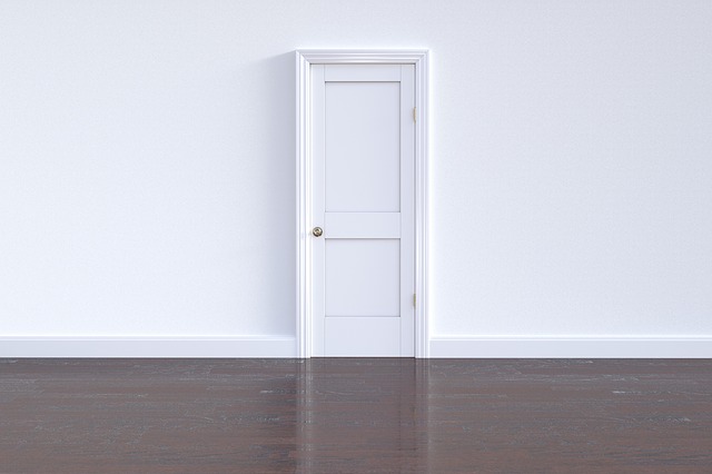 Looking over a dark brown wood floor at a white will with a white door.